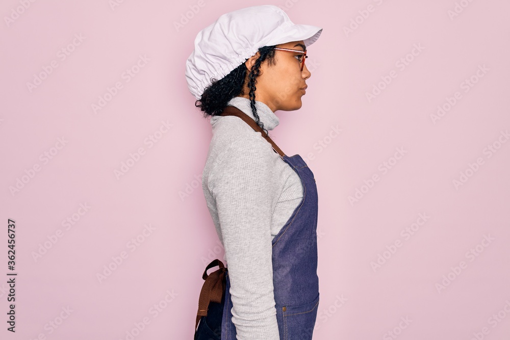 Young african american curly baker woman wearing apron and cap over pink background looking to side, relax profile pose with natural face with confident smile. Young african american curly baker woman wearing apron and cap over pink background looking to side, relax profile pose with natural face with confident smile.