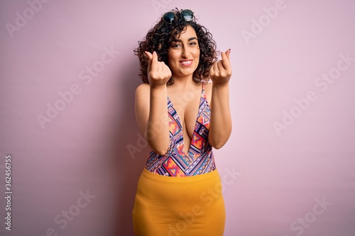 Young beautiful arab woman on vacation wearing swimsuit and sunglasses over pink background doing money gesture with hands, asking for salary payment, millionaire business