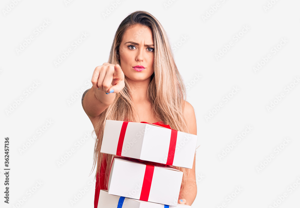 Young beautiful blonde woman holding birthday gifts pointing with finger to the camera and to you, confident gesture looking serious Young beautiful blonde woman holding birthday gifts pointing with finger to the camera and to you, confident gesture looking serious