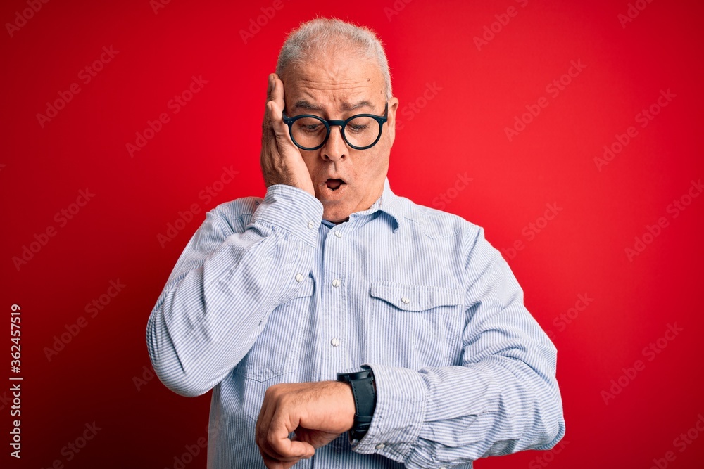 Middle age handsome hoary man wearing casual striped shirt and glasses over red background Looking at the watch time worried, afraid of getting late Middle age handsome hoary man wearing casual striped shirt and glasses over red background Looking at the watch time worried, afraid of getting late