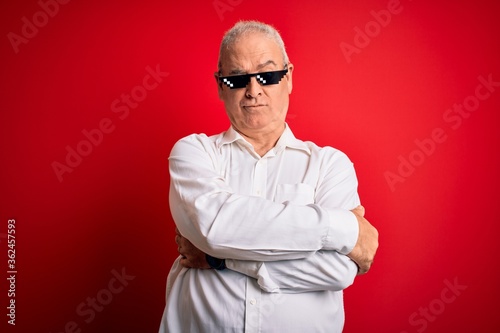 Middle age hoary man wearing funny sunglasses over isolated red background skeptic and nervous, disapproving expression on face with crossed arms. Negative person.