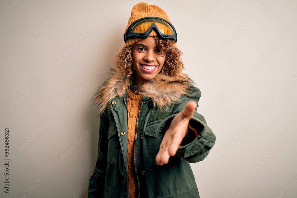 Young african american skier woman with curly hair wearing snow sportswear and ski goggles smiling cheerful offering palm hand giving assistance and acceptance.