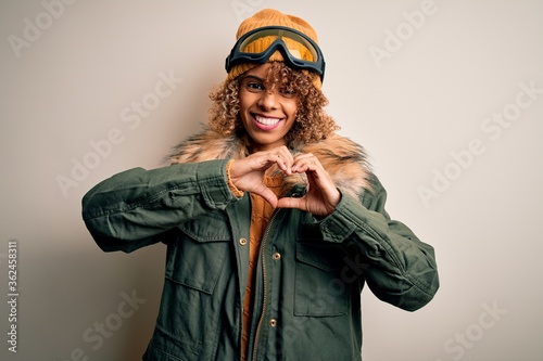 Young african american skier woman with curly hair wearing snow sportswear and ski goggles smiling in love doing heart symbol shape with hands. Romantic concept.