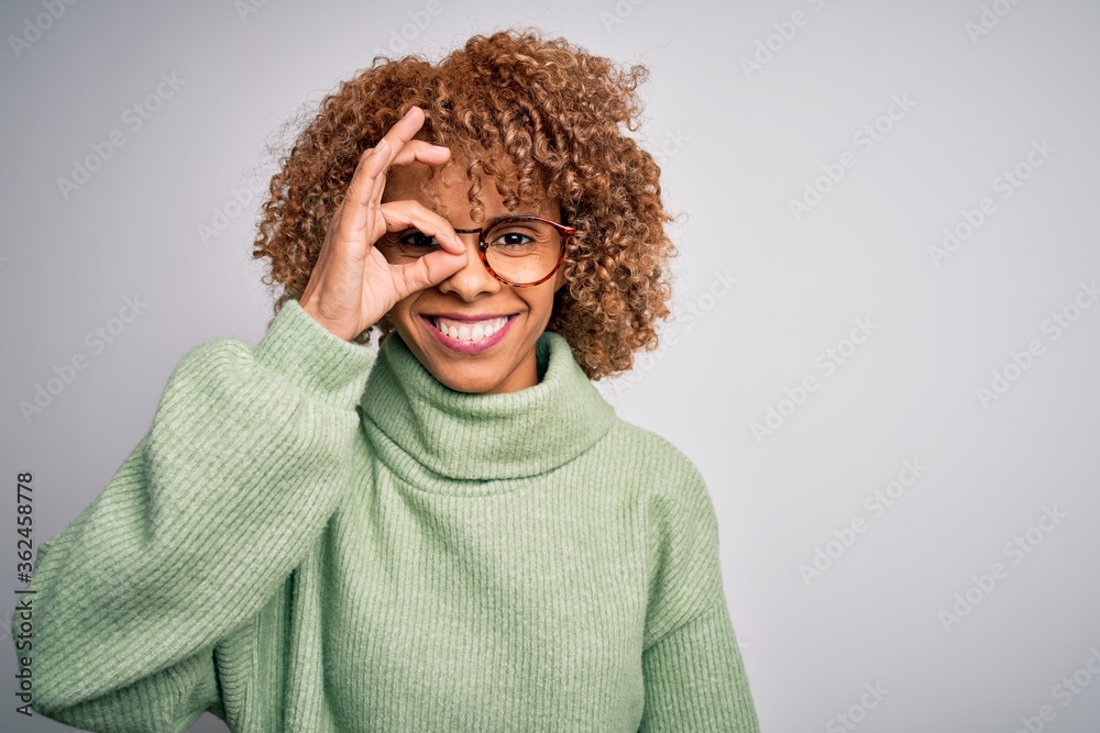 Young beautiful african american woman wearing turtleneck sweater and glasses doing ok gesture with hand smiling, eye looking through fingers with happy face. Young beautiful african american woman wearing turtleneck sweater and glasses doing ok gesture with hand smiling, eye looking through fingers with happy face.