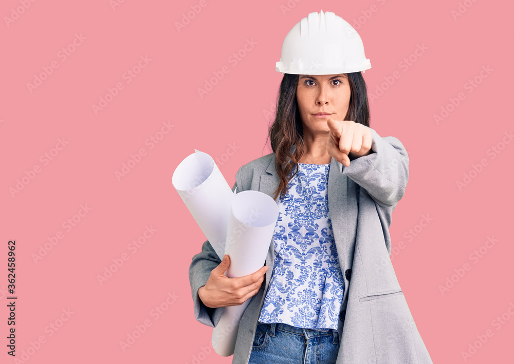 Young beautiful brunette woman wearing architect hardhat holding blueprint pointing with finger to the camera and to you, confident gesture looking serious Young beautiful brunette woman wearing architect hardhat holding blueprint pointing with finger to the camera and to you, confident gesture looking serious