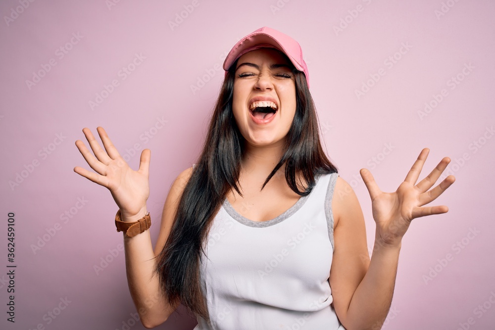 Young brunette woman wearing casual sport cap over pink background celebrating mad and crazy for success with arms raised and closed eyes screaming excited. Winner concept Young brunette woman wearing casual sport cap over pink background celebrating mad and crazy for success with arms raised and closed eyes screaming excited. Winner concept