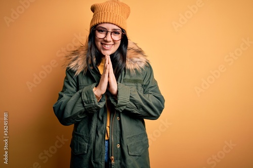 Young brunette woman wearing glasses and winter coat with hat over yellow isolated background praying with hands together asking for forgiveness smiling confident. © Krakenimages.com