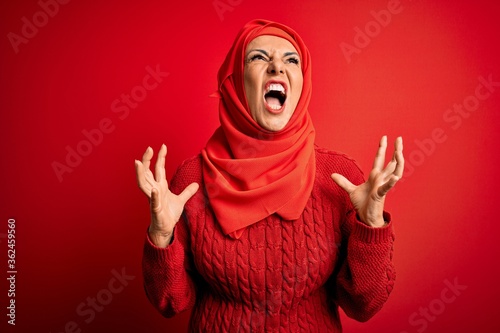 Middle age woman wearing traditional muslim hijab standing over isolated red background crazy and mad shouting and yelling with aggressive expression and arms raised. Frustration concept.