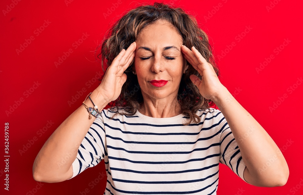 Middle age beautiful brunette woman wearing striped t-shirt standing over red background with hand on head, headache because stress. Suffering migraine. Middle age beautiful brunette woman wearing striped t-shirt standing over red background with hand on head, headache because stress. Suffering migraine.