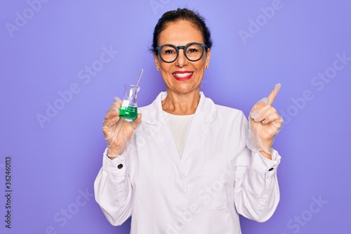 Middle age senior scientist woman wearing laboratory coat holding research test tube very happy pointing with hand and finger to the side