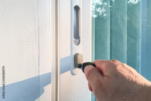 Closeup on male hand puts the key in the keyhole to open exterior white vinyl PVC entrance door with glass and reflection on summer twilight tropical season.