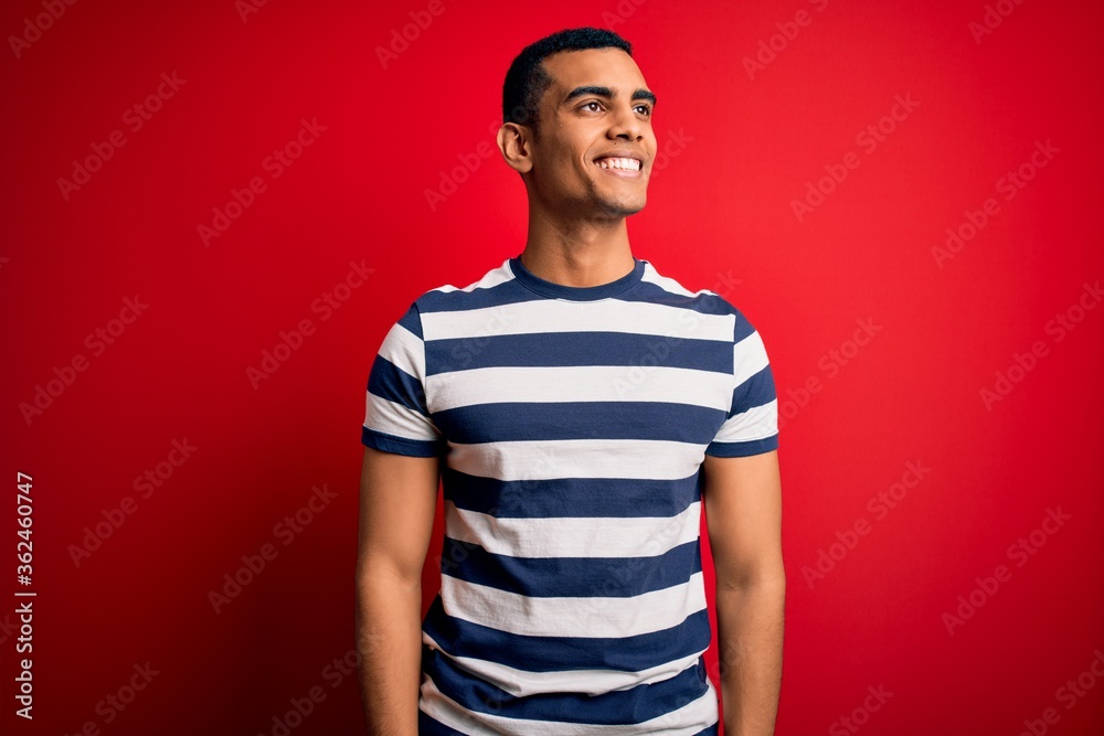 Handsome african american man wearing casual striped t-shirt standing over red background looking away to side with smile on face, natural expression. Laughing confident. Handsome african american man wearing casual striped t-shirt standing over red background looking away to side with smile on face, natural expression. Laughing confident.
