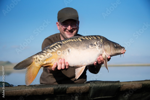 Happy smiling fisherman holding his fish with lake on the background. Selective focus on the carp.
