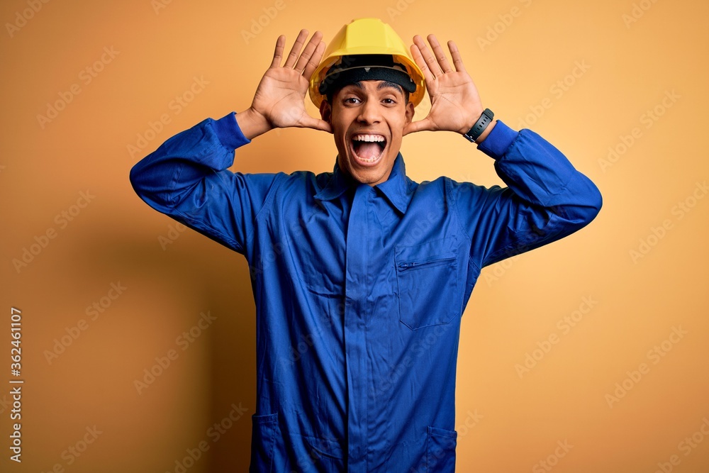 Young handsome african american worker man wearing blue uniform and security helmet Smiling cheerful playing peek a boo with hands showing face. Surprised and exited