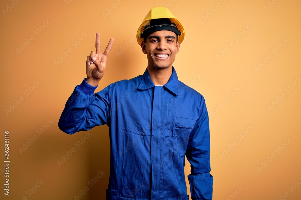Young handsome african american worker man wearing blue uniform and security helmet smiling looking to the camera showing fingers doing victory sign. Number two.