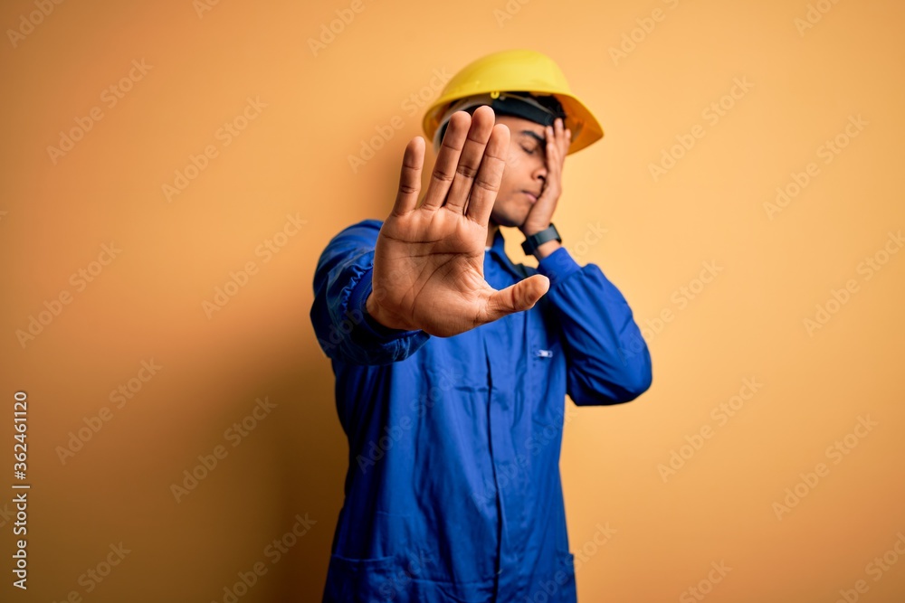 Young handsome african american worker man wearing blue uniform and security helmet covering eyes with hands and doing stop gesture with sad and fear expression. Embarrassed and negative concept.