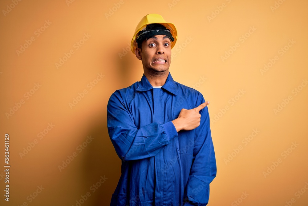 Young handsome african american worker man wearing blue uniform and security helmet Pointing aside worried and nervous with forefinger, concerned and surprised expression