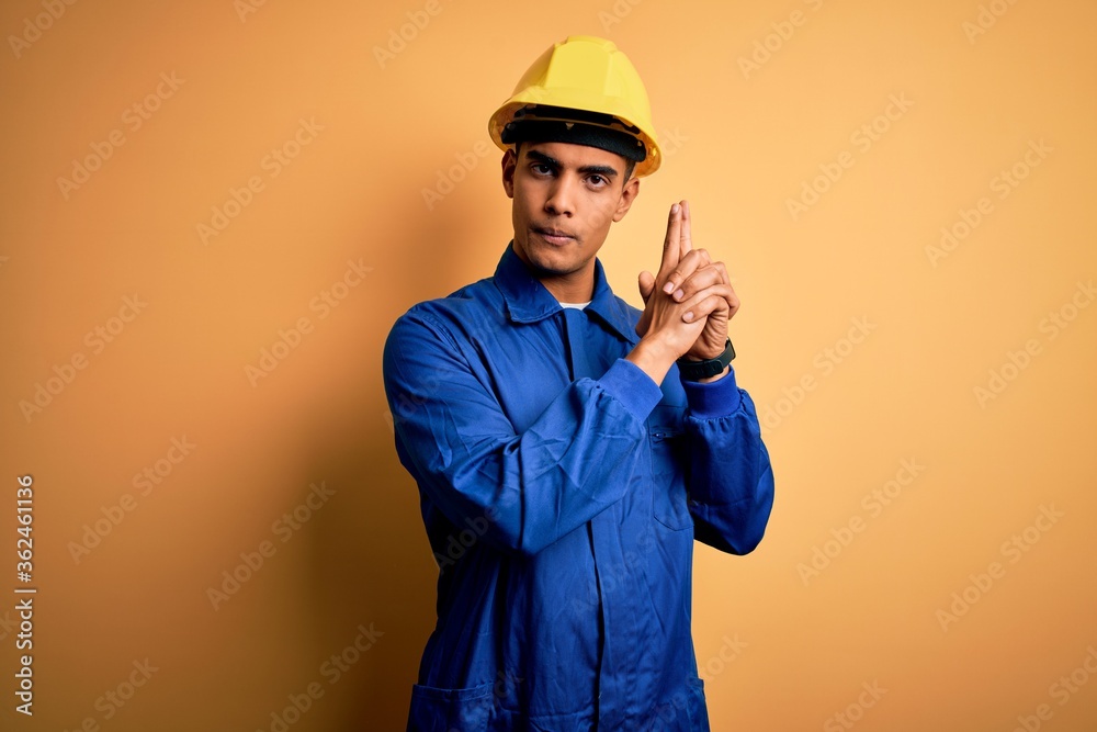 Young handsome african american worker man wearing blue uniform and security helmet Holding symbolic gun with hand gesture, playing killing shooting weapons, angry face Young handsome african american worker man wearing blue uniform and security helmet Holding symbolic gun with hand gesture, playing killing shooting weapons, angry face