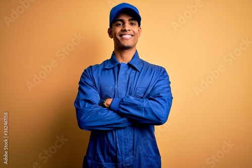 Young african american mechanic man wearing blue uniform and cap over yellow background happy face smiling with crossed arms looking at the camera. Positive person.
