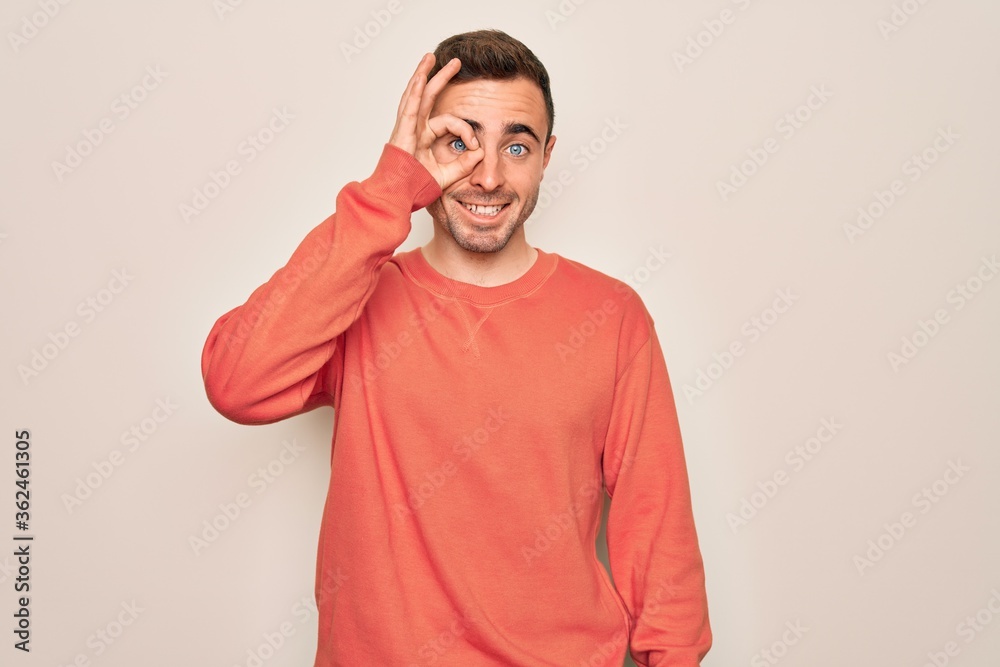 Young handsome man with blue eyes wearing casual sweater standing over white background doing ok gesture with hand smiling, eye looking through fingers with happy face. Young handsome man with blue eyes wearing casual sweater standing over white background doing ok gesture with hand smiling, eye looking through fingers with happy face.