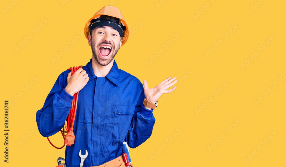 Young hispanic man wearing elecrician uniform holding cable celebrating victory with happy smile and winner expression with raised hands Young hispanic man wearing elecrician uniform holding cable celebrating victory with happy smile and winner expression with raised hands