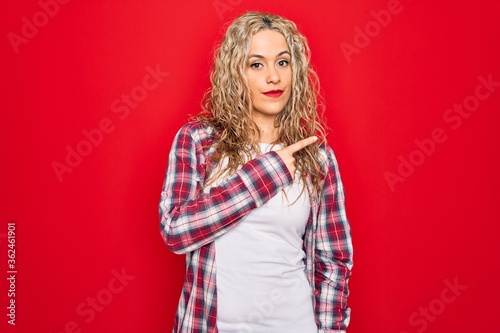 Young beautiful blonde woman wearing casual shirt standing over isolated red background Pointing with hand finger to the side showing advertisement, serious and calm face