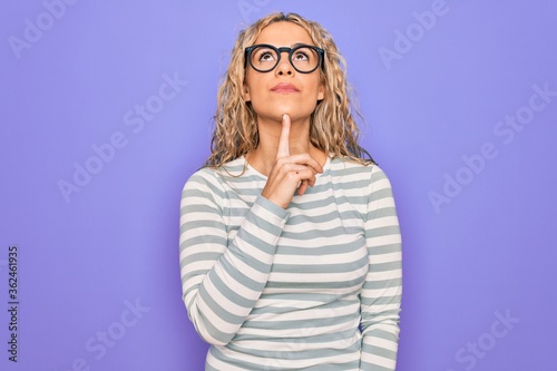 Beautiful blonde woman wearing casual striped t-shirt and glasses over purple background Thinking concentrated about doubt with finger on chin and looking up wondering