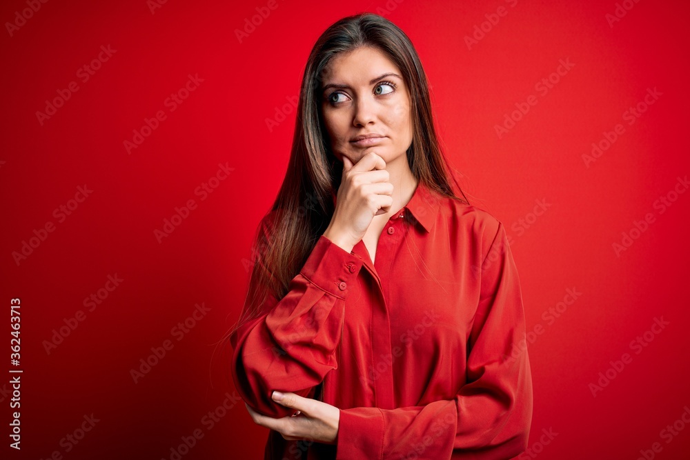 Young beautiful woman with blue eyes wearing casual shirt standing over red background with hand on chin thinking about question, pensive expression. Smiling with thoughtful face. Doubt concept.