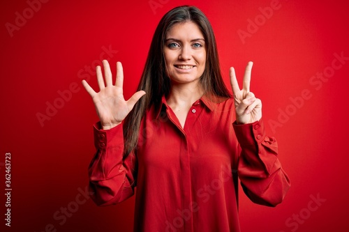 Young beautiful woman with blue eyes wearing casual shirt standing over red background showing and pointing up with fingers number seven while smiling confident and happy. © Krakenimages.com