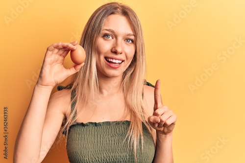 Young blonde woman holding egg smiling with an idea or question pointing finger with happy face, number one