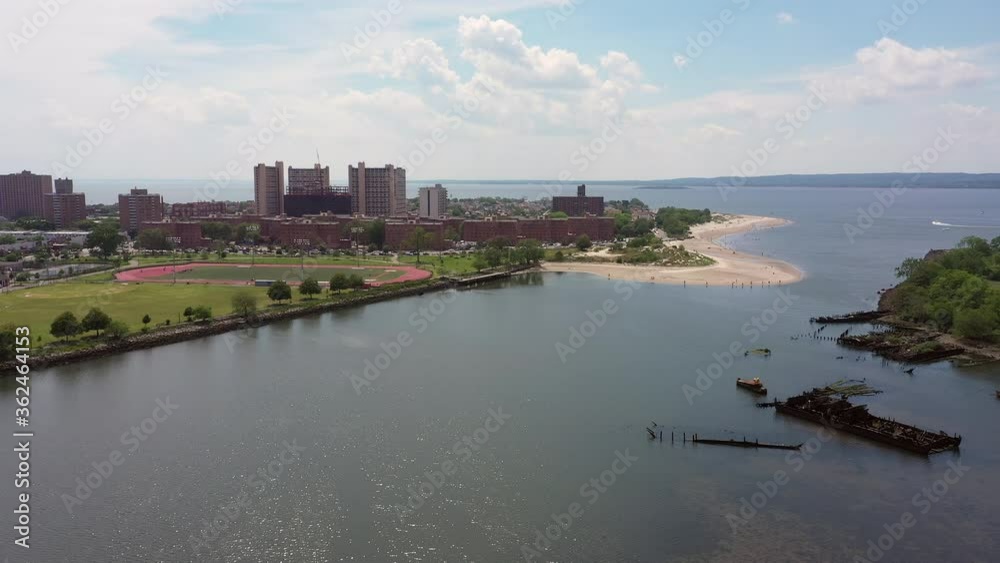 Drone flies over a bayside park with a sandy tanned beach with large brown buildings in the distance.