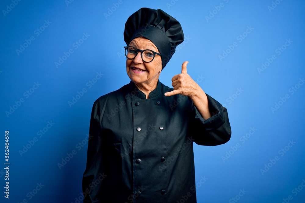 Senior beautiful grey-haired chef woman wearing cooker uniform and hat over blue background smiling doing phone gesture with hand and fingers like talking on the telephone. Communicating concepts.