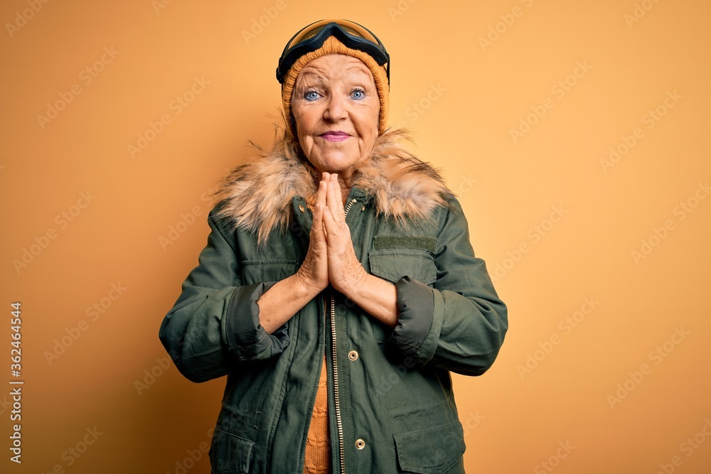 Senior beautiful grey-haired skier woman wearing snow sportswear and ski goggles praying with hands together asking for forgiveness smiling confident. Senior beautiful grey-haired skier woman wearing snow sportswear and ski goggles praying with hands together asking for forgiveness smiling confident.