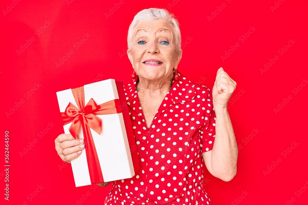 Senior beautiful woman with blue eyes and grey hair holding gift screaming proud, celebrating victory and success very excited with raised arm Senior beautiful woman with blue eyes and grey hair holding gift screaming proud, celebrating victory and success very excited with raised arm