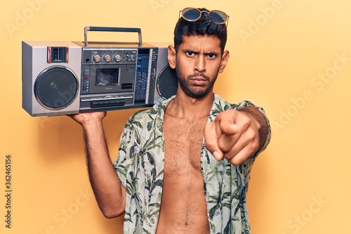 Young latin man wearing summer shirt holding boombox pointing with finger to the camera and to you, confident gesture looking serious