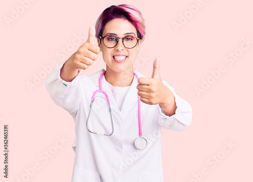 Young beautiful woman with pink hair wearing doctor uniform approving doing positive gesture with hand  thumbs up smiling and happy for success. winner gesture.