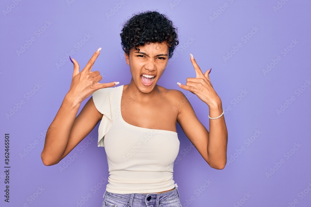 Young beautiful african american afro woman wearing casual t-shirt over purple background shouting with crazy expression doing rock symbol with hands up. Music star. Heavy music concept. Young beautiful african american afro woman wearing casual t-shirt over purple background shouting with crazy expression doing rock symbol with hands up. Music star. Heavy music concept.