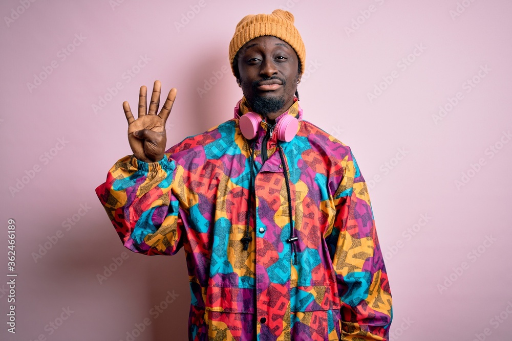 Young handsome african american man wearing colorful coat and cap over pink background showing and pointing up with fingers number four while smiling confident and happy.