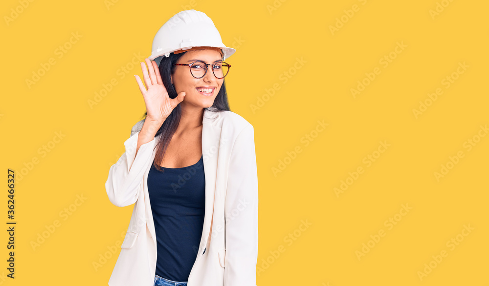 Young beautiful latin girl wearing architect hardhat and glasses smiling with hand over ear listening an hearing to rumor or gossip. deafness concept.