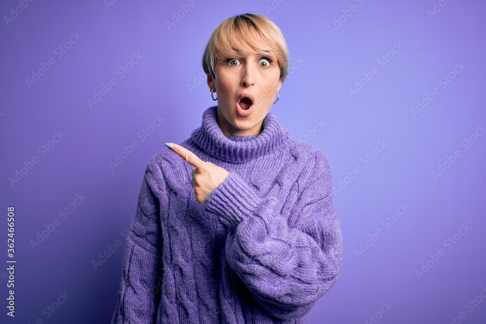 Young blonde woman with short hair wearing winter turtleneck sweater over purple background Surprised pointing with finger to the side, open mouth amazed expression. Young blonde woman with short hair wearing winter turtleneck sweater over purple background Surprised pointing with finger to the side, open mouth amazed expression.