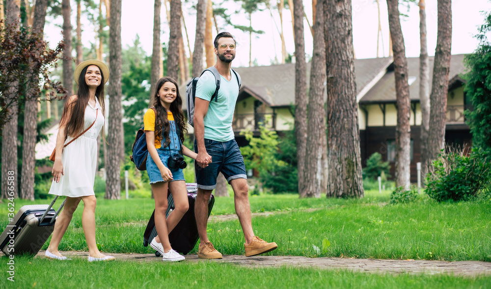 Family vacation in the hotel in the forest. A full-length photo of the young parents, and their teenage daughter, walking to the hotel in the forest, with their suitcases and backpacks.