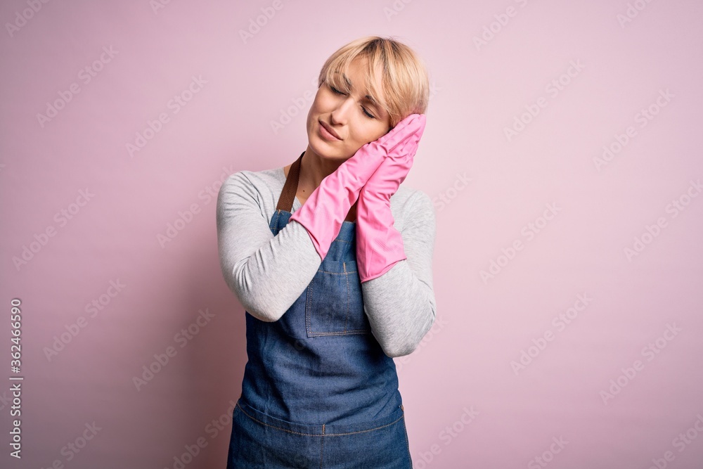 Young blonde cleaner woman with short hair wearing apron and gloves over pink background sleeping tired dreaming and posing with hands together while smiling with closed eyes.