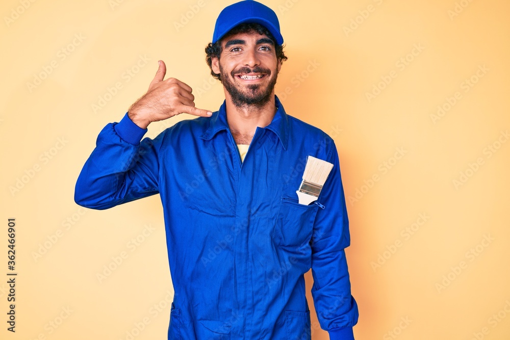 Handsome young man with curly hair and bear wearing builder jumpsuit uniform smiling doing phone gesture with hand and fingers like talking on the telephone. communicating concepts.