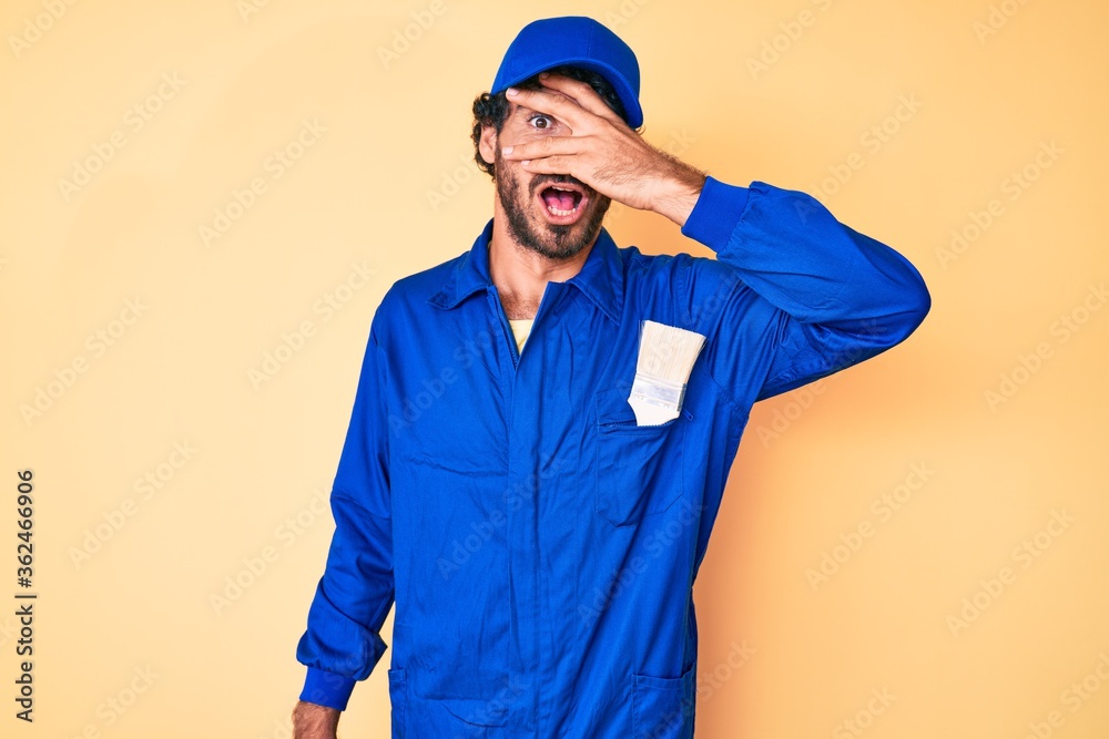 Handsome young man with curly hair and bear wearing builder jumpsuit uniform peeking in shock covering face and eyes with hand, looking through fingers with embarrassed expression.