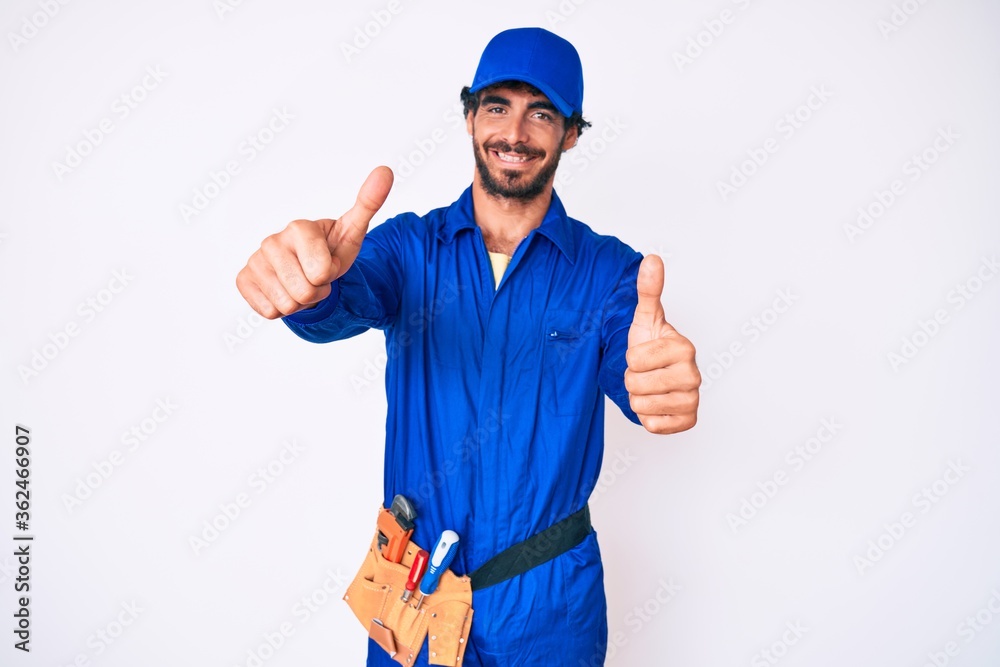 Handsome young man with curly hair and bear weaing handyman uniform approving doing positive gesture with hand, thumbs up smiling and happy for success. winner gesture.