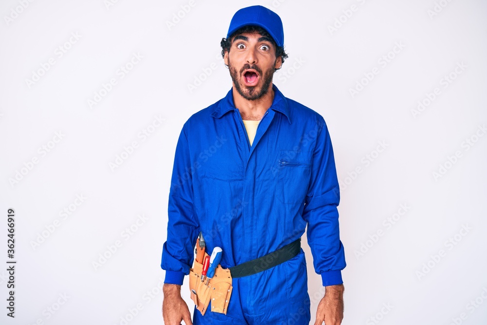 Handsome young man with curly hair and bear weaing handyman uniform afraid and shocked with surprise and amazed expression, fear and excited face.