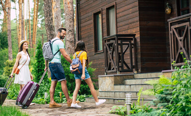 Nice hotel. A photo of a happy family with luggages and backpacs on a weekend coming to the wooden hotel surrounded by trees, father holding his daughter's hand.