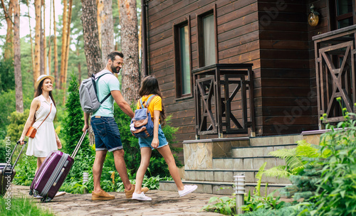 Nice hotel. A photo of a happy family with luggages and backpacs on a weekend coming to the wooden hotel surrounded by trees, father holding his daughter's hand.