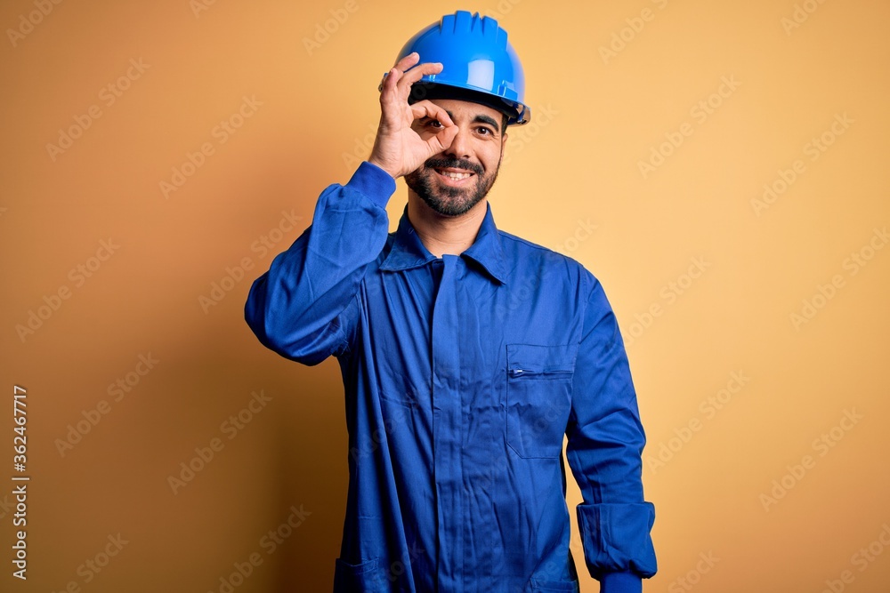 Mechanic man with beard wearing blue uniform and safety helmet over yellow background doing ok gesture with hand smiling, eye looking through fingers with happy face.