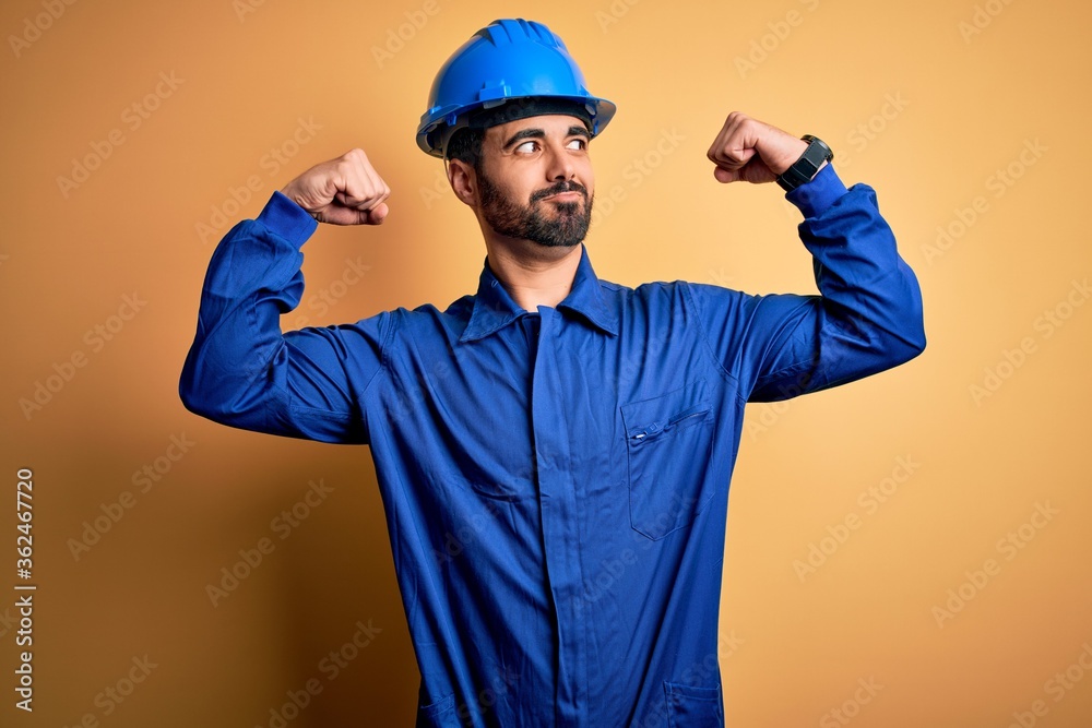 Mechanic man with beard wearing blue uniform and safety helmet over yellow background showing arms muscles smiling proud. Fitness concept.